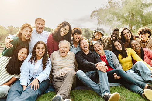 Group of 15 people of diverse ages and races sitting on the ground and smiling at the camera.