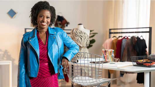  Joven negra diseñadora de ropa posa en su estudio sonriendo. Lleva una chaqueta de color azul intenso.