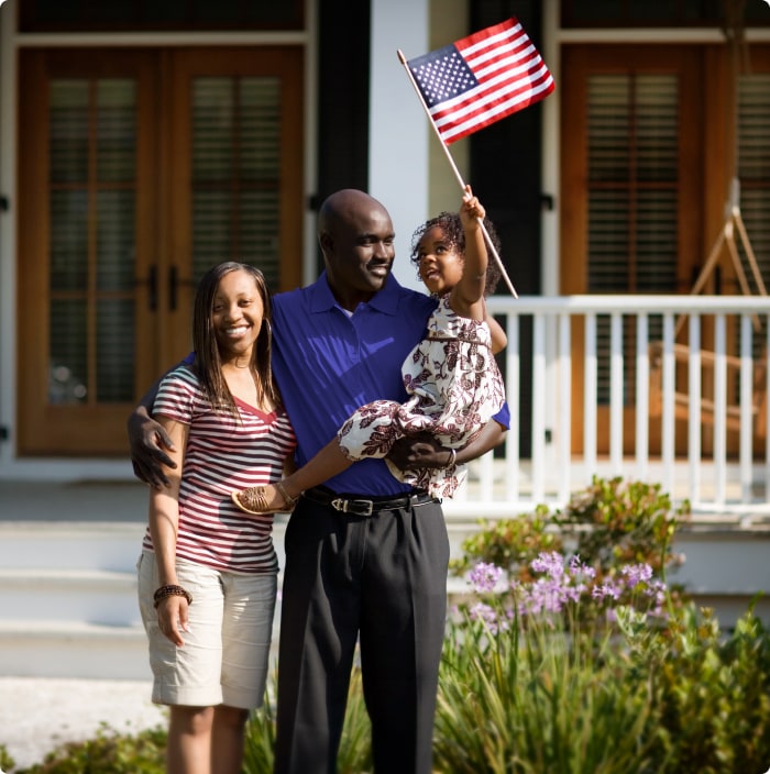 A dad holding a little girl waving an American flag and mom pose, smiling, in front of a house