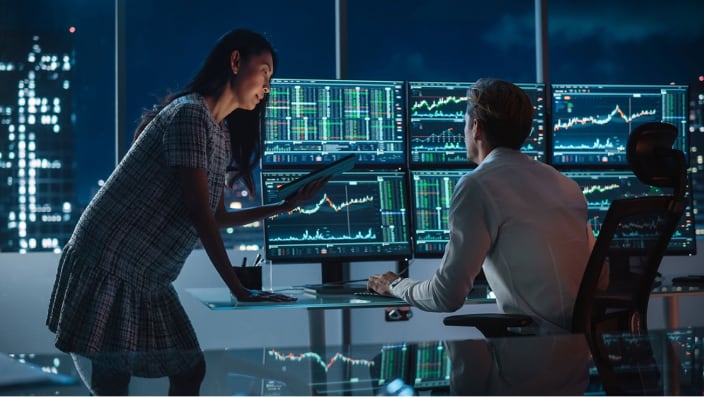 Professional man and woman working at a large desk looking at six computer monitors