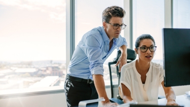 One person standing with another person sitting in front of a computer while both look at the monitor.