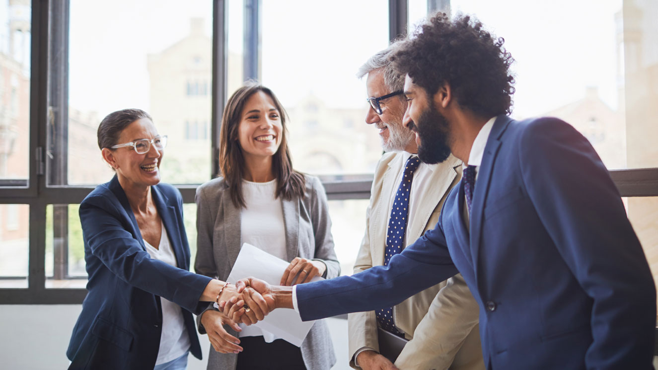 Two men and two women in professional suits. One man and woman are shaking hands while the other two look on. The group is discussing corporate trust solutions.