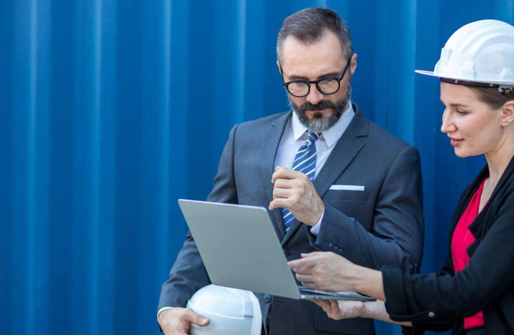 Man in a suit standing next to a woman in hardhat and she is pointing at a laptop showing him something on the screen.