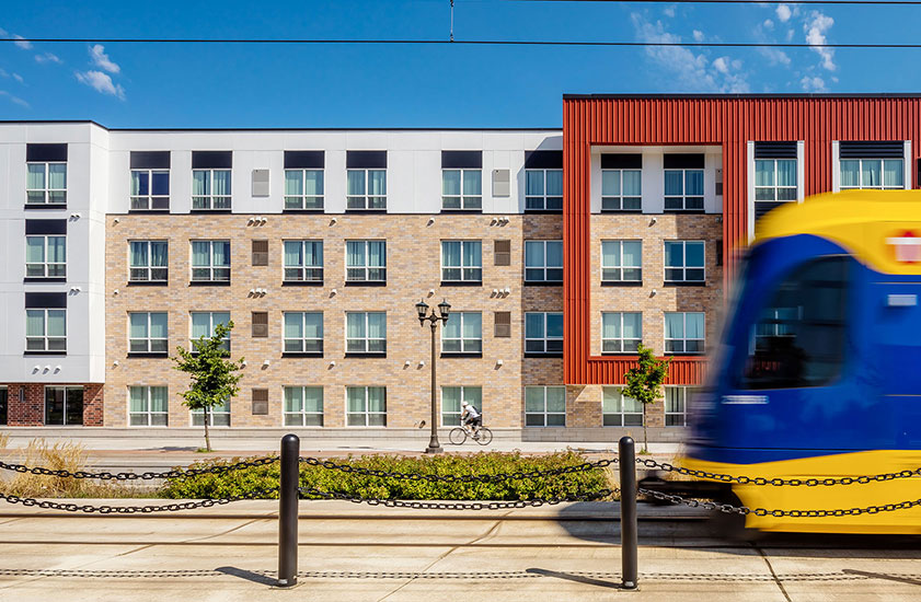 Light-rail train passing in front of a four-story apartment complex.