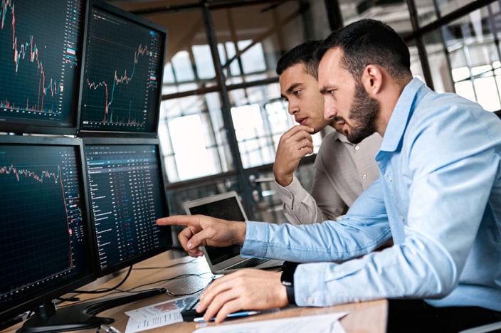 Two male coworkers looking at four large monitors showing financial charts