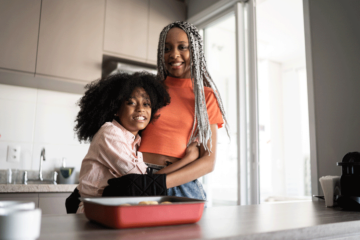 African-American mom and daughter standing in the kitchen of their home and smiling at the camera.