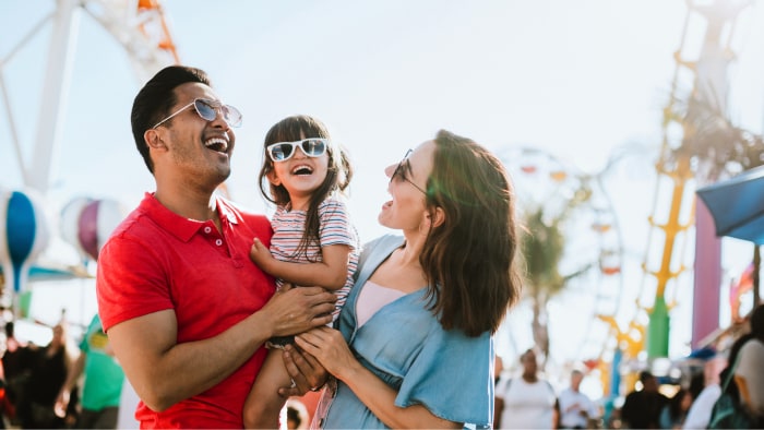 Una familia hispana que incluye a la mamá, al papá y a una pequeña niña sonríen y ríen juntos en un carnaval.