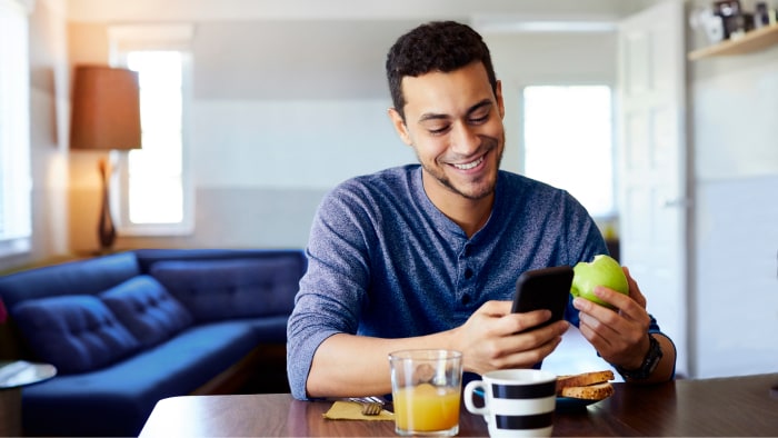 Un joven hispano viendo su teléfono y sonriendo mientras desayuna.